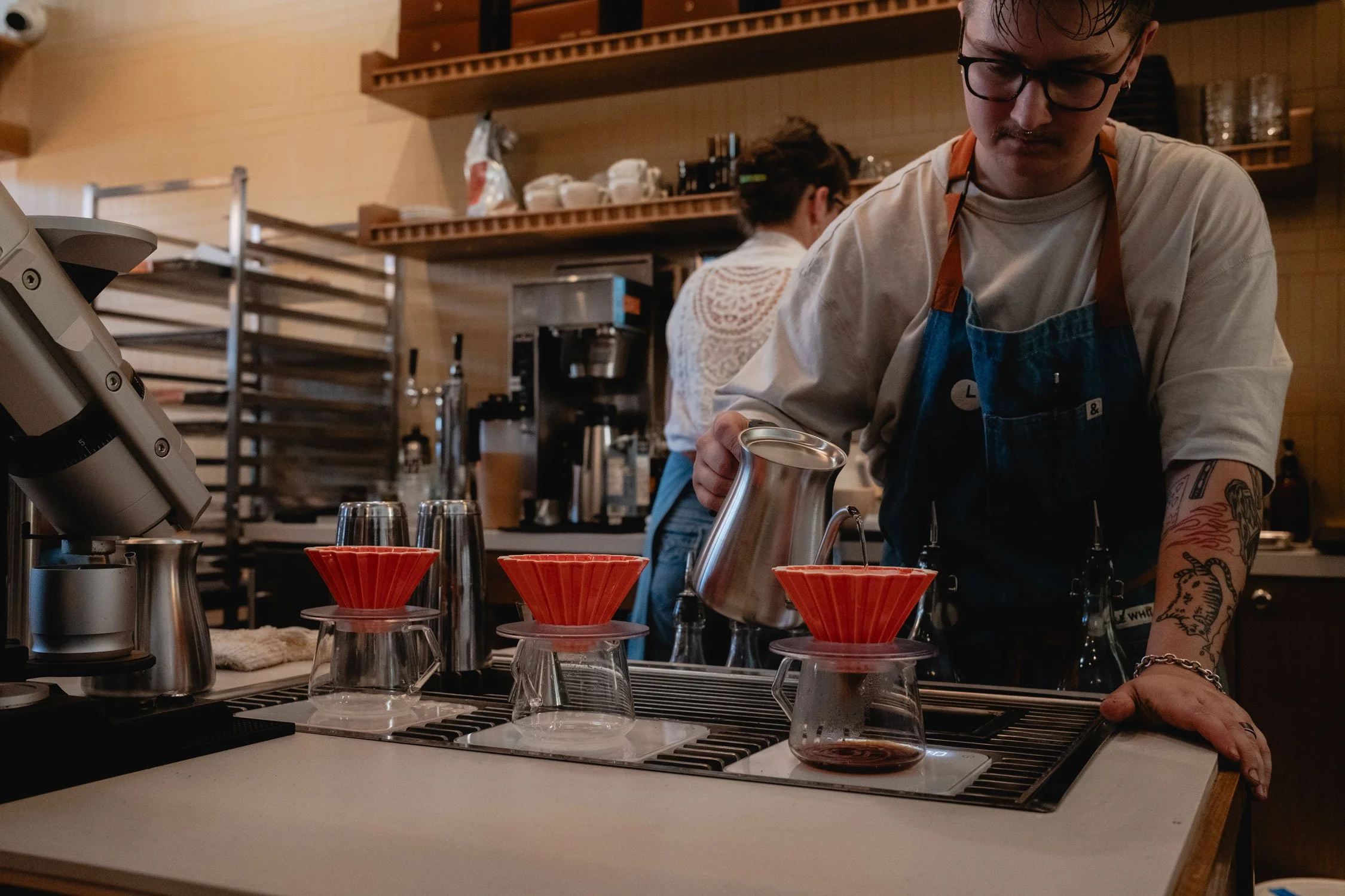 Barista pouring hot water into coffee drippers placed on upside-down glasses at coffee bar.