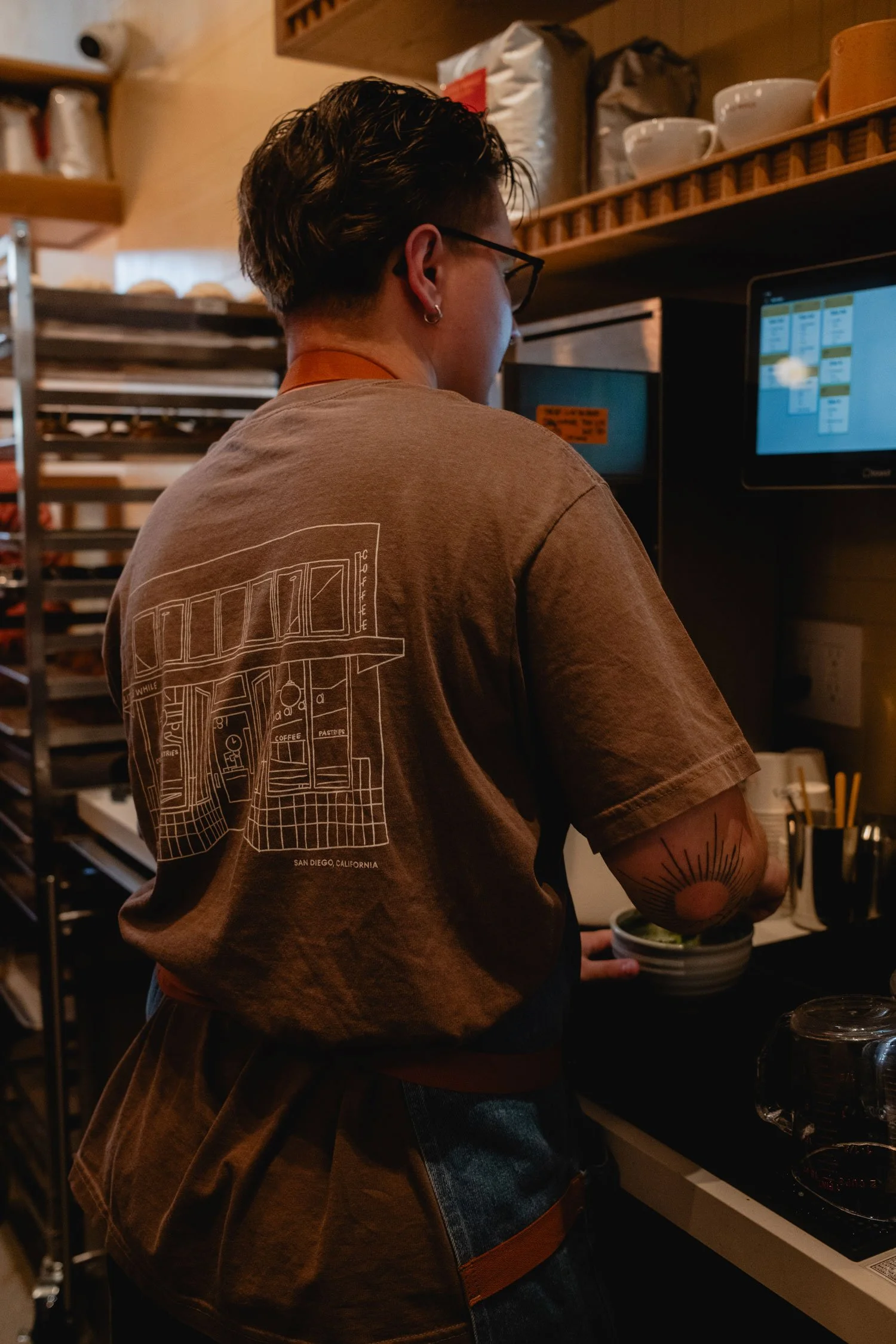 A person with short dark hair, glasses, and a tattoo on their arm, working behind a coffee shop counter, preparing a beverage.