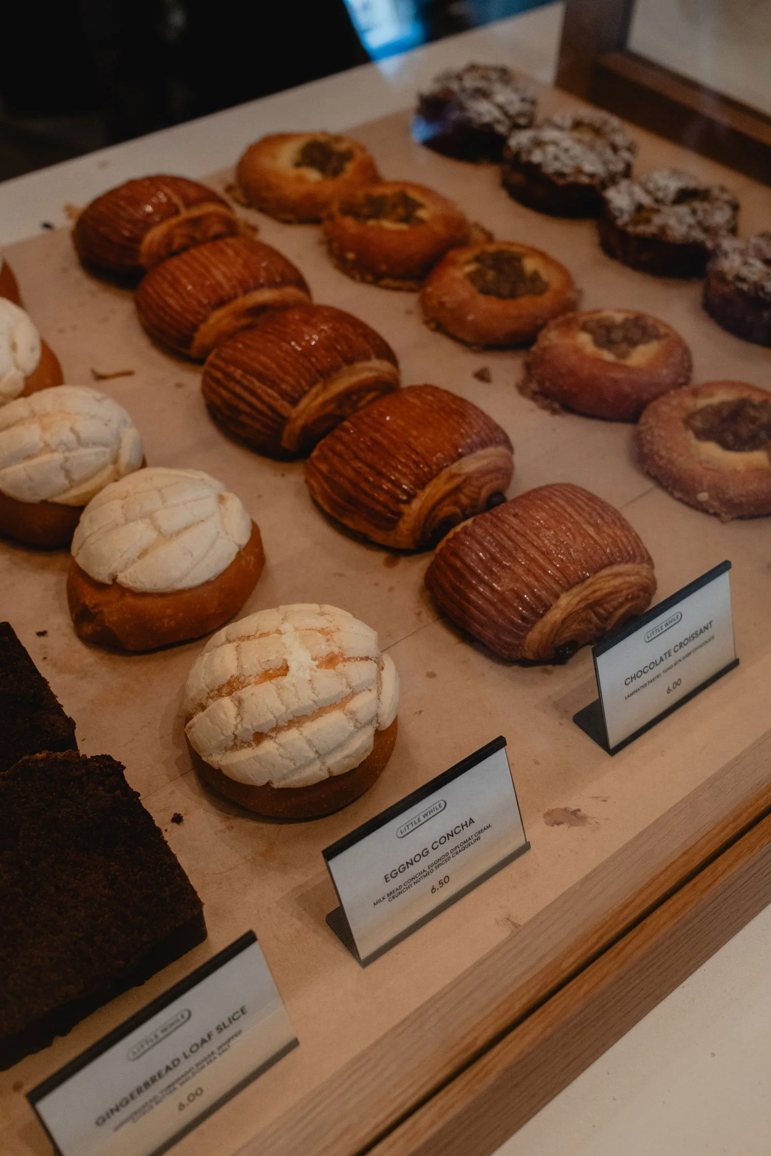 Display of baked goods on a tray, including eggnog conchas, chocolate croissants, and other pastries, with small price tags