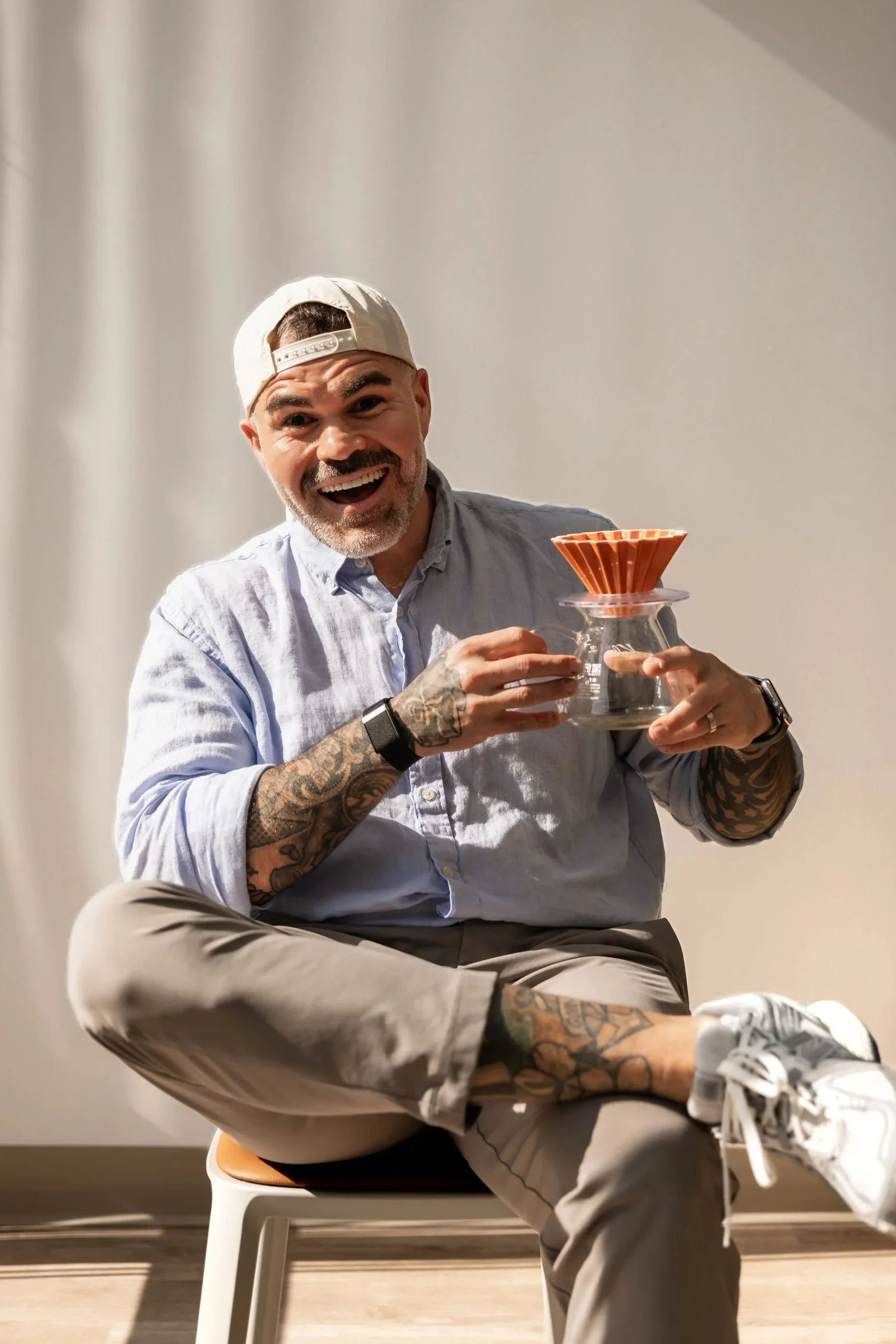 A man with a beard and tattoos, wearing a backwards cap and a light blue button-up shirt, is sitting cross-legged on a chair. He is smiling and holding a Chemex coffee maker with a paper filter inside. The background is plain and light-colored.