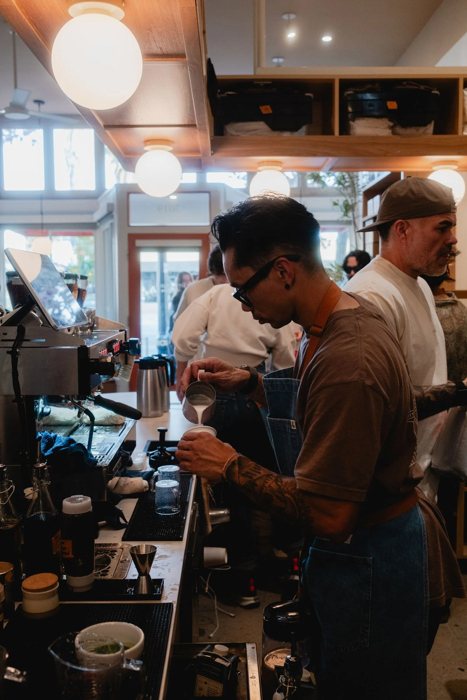 Barista pouring milk into a cup at a busy coffee shop with customers in the background.