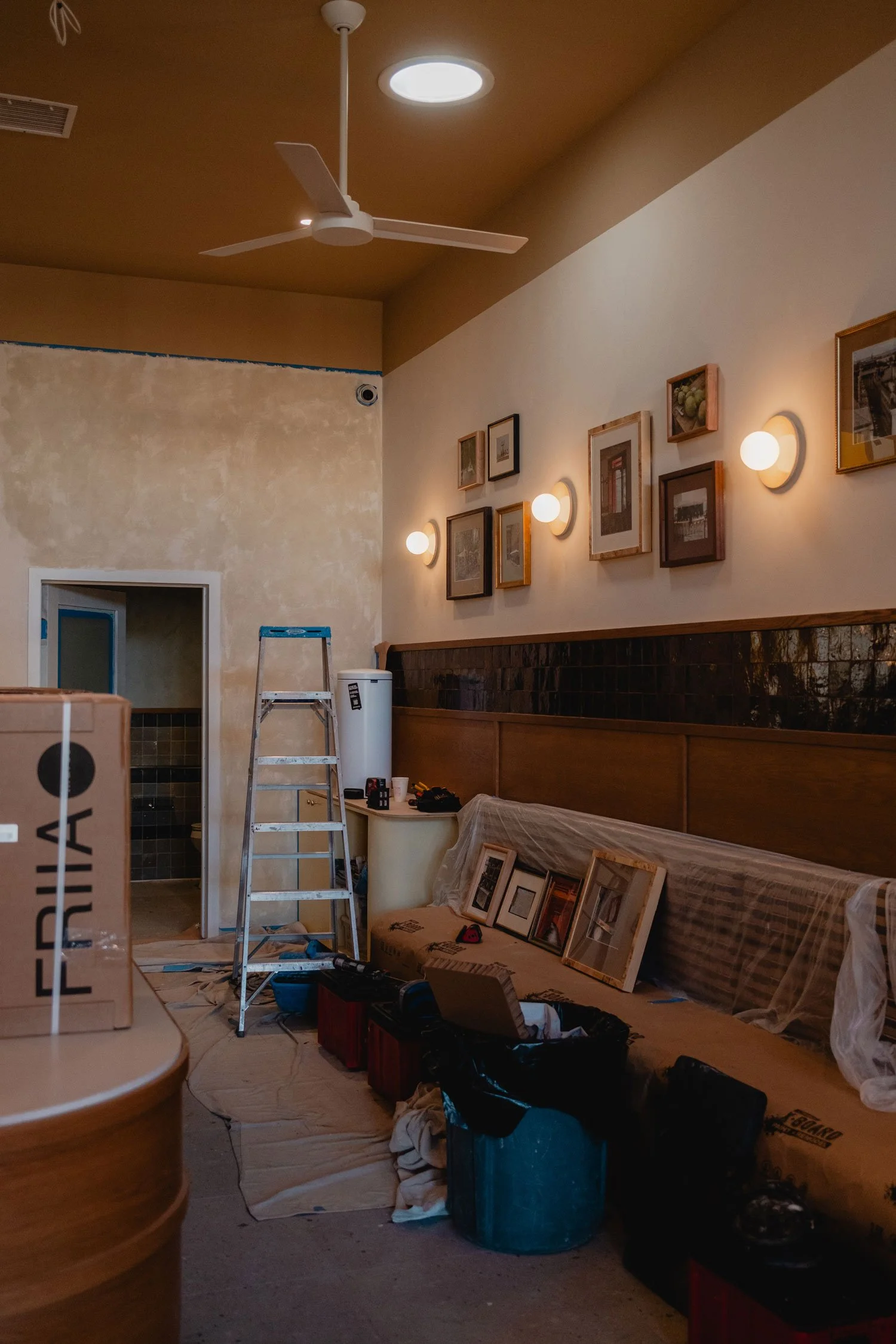 A room under construction or renovation with a ladder, framed pictures, a ladder, wall-mounted lights, and various tools and materials scattered on the floor.
