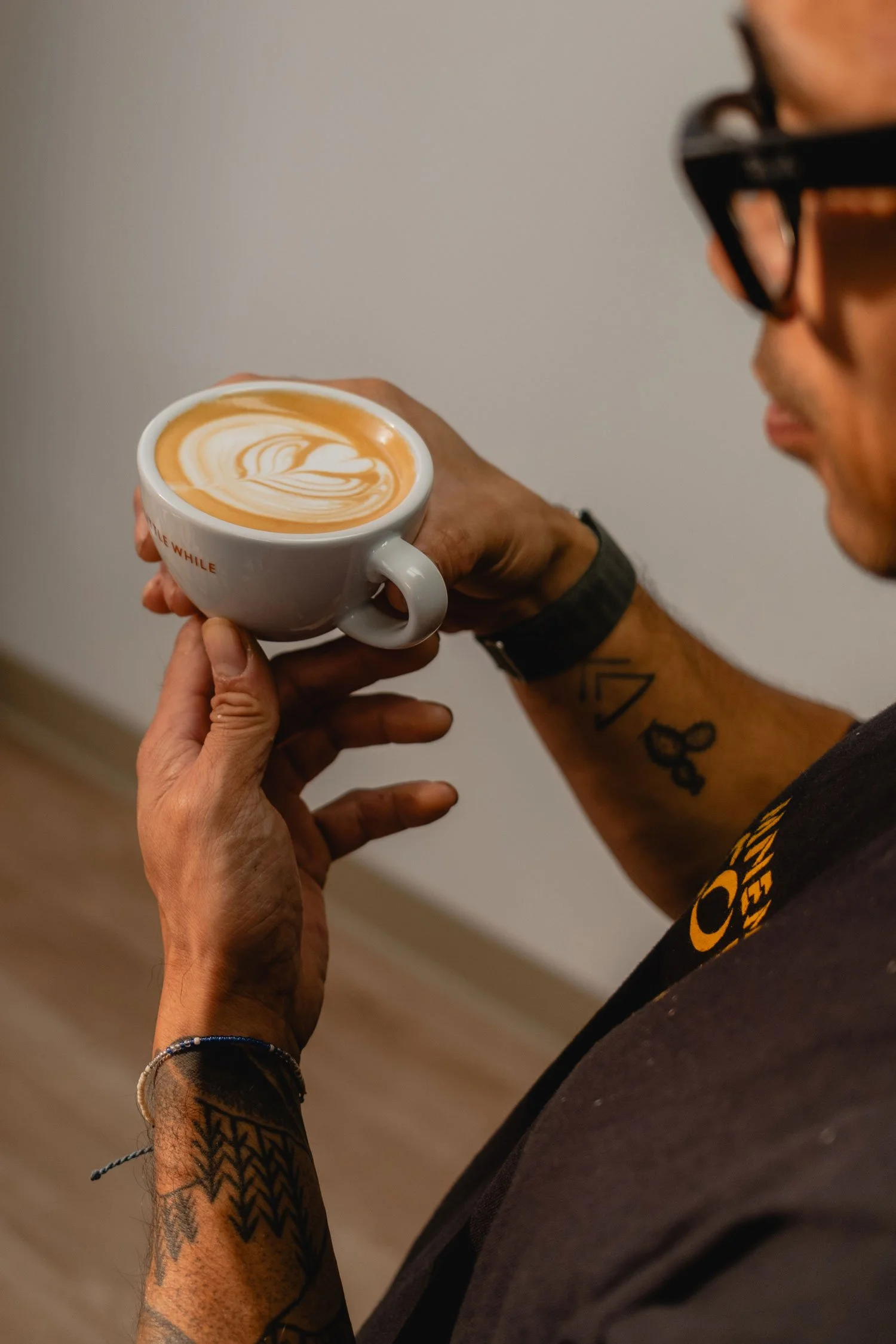 A man with tattoos on his arms and neck, wearing glasses, holds a coffee cup with latte art in a white mug.