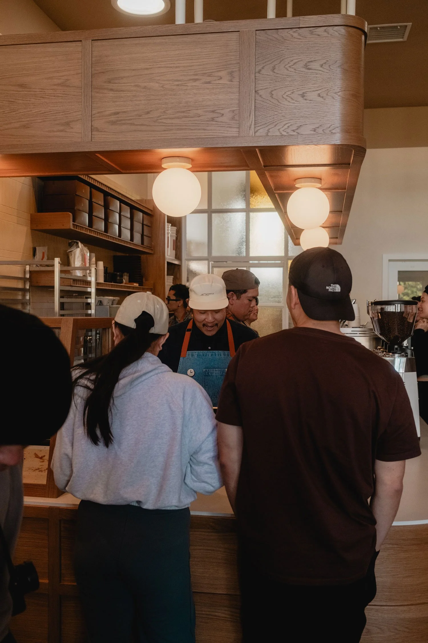 Group of people inside a coffee shop, ordering or waiting for their drinks, with baristas preparing beverages behind the counter.