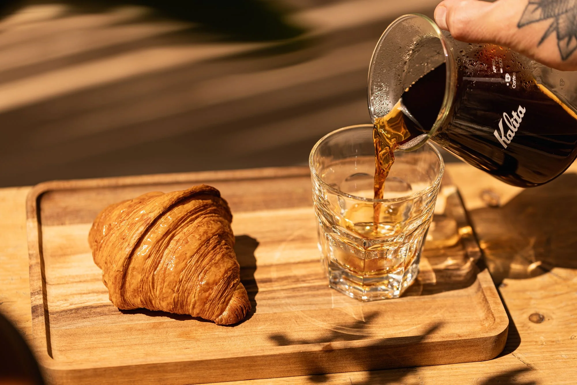 A croissant on a wooden tray with a glass of coffee being filled from a pour-over coffee pot.