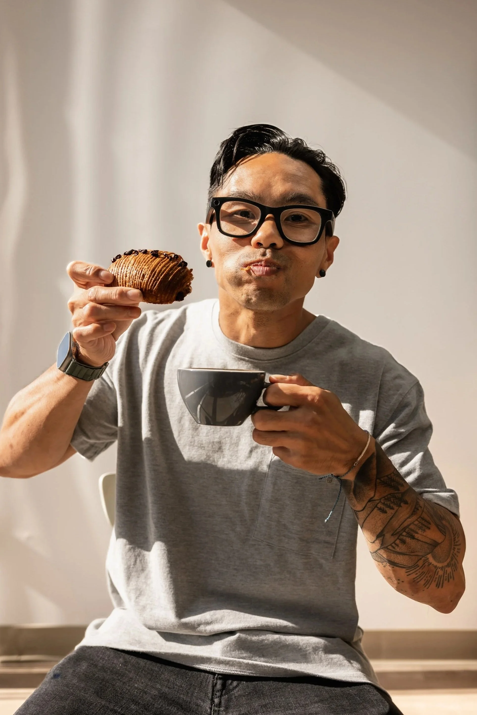 Man with glasses and tattoos enjoying a coffee and a pastry at home.