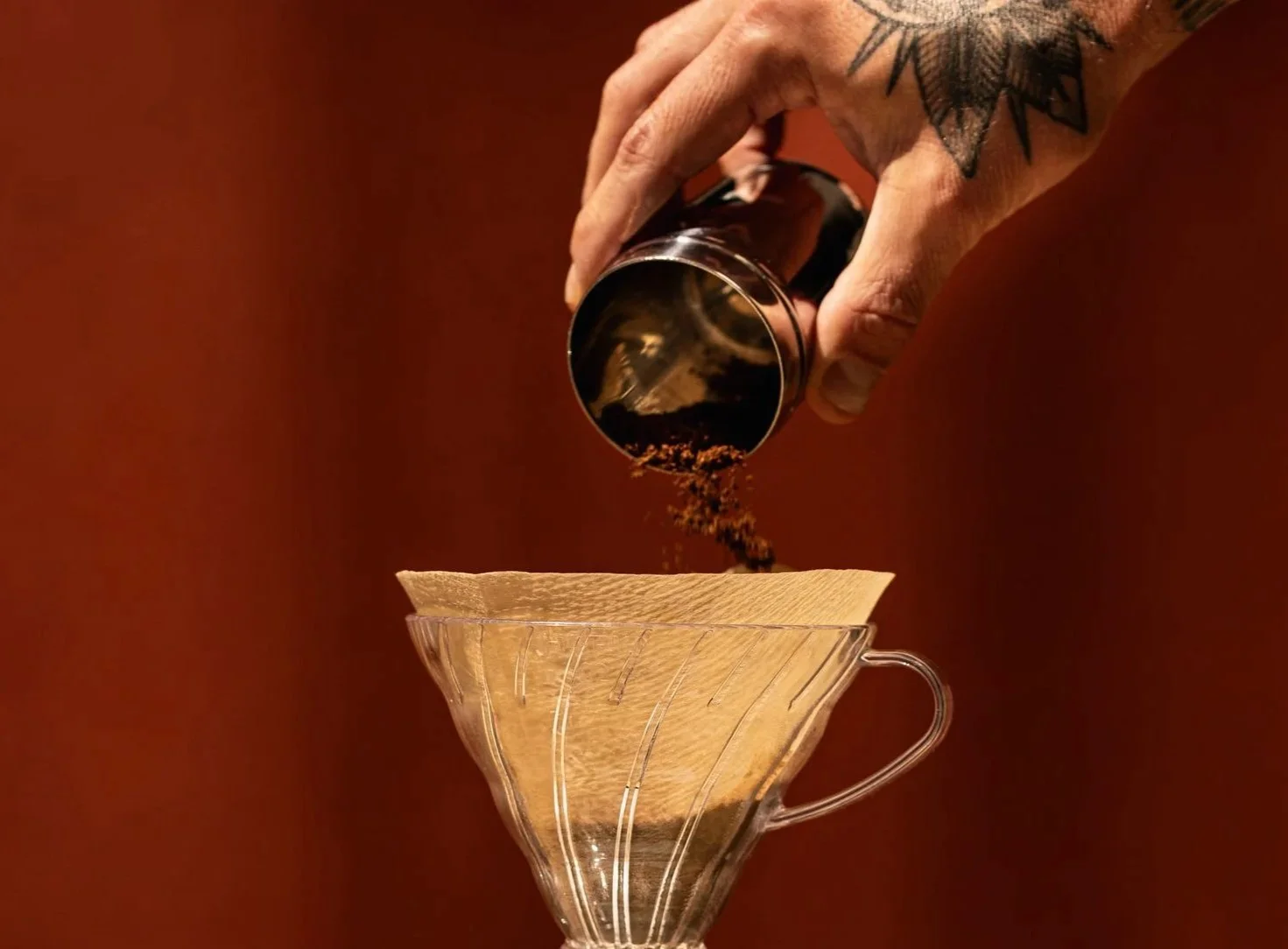 Hand with a tattoo pouring ground coffee into a glass coffee dripper.