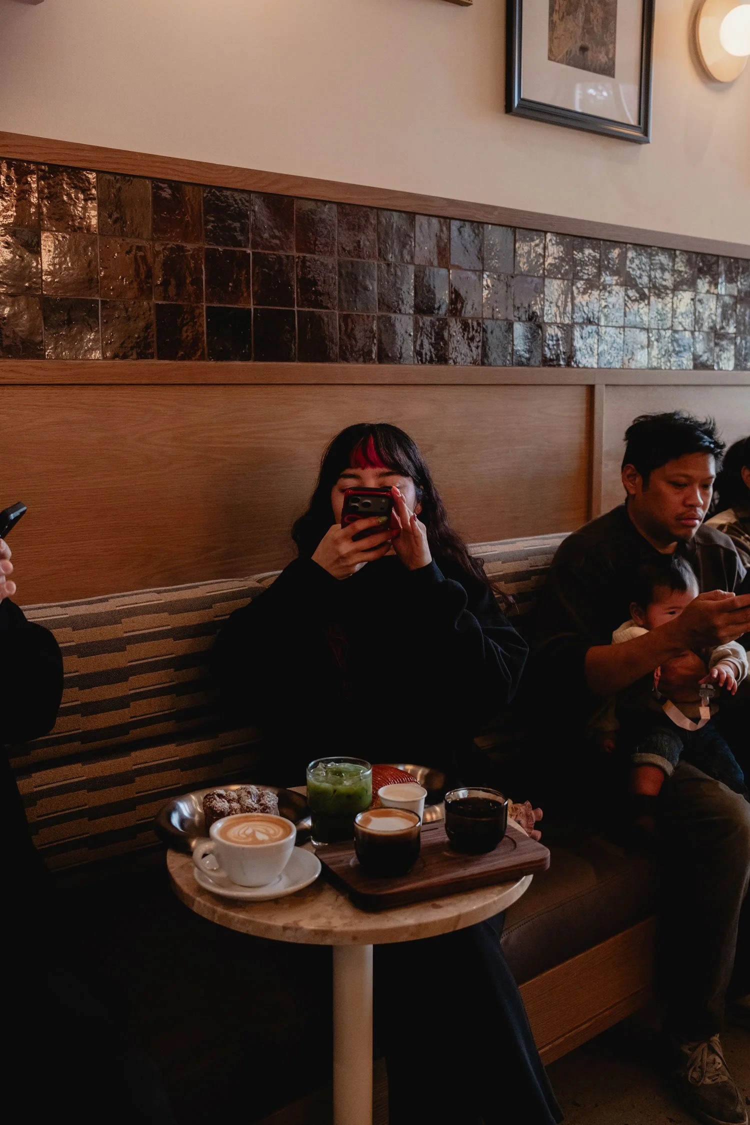 A woman with dark hair and red highlights takes a photo with her phone inside a cafe. On the table in front of her are coffee drinks, a green iced beverage, a dessert, and a wooden tray. 
