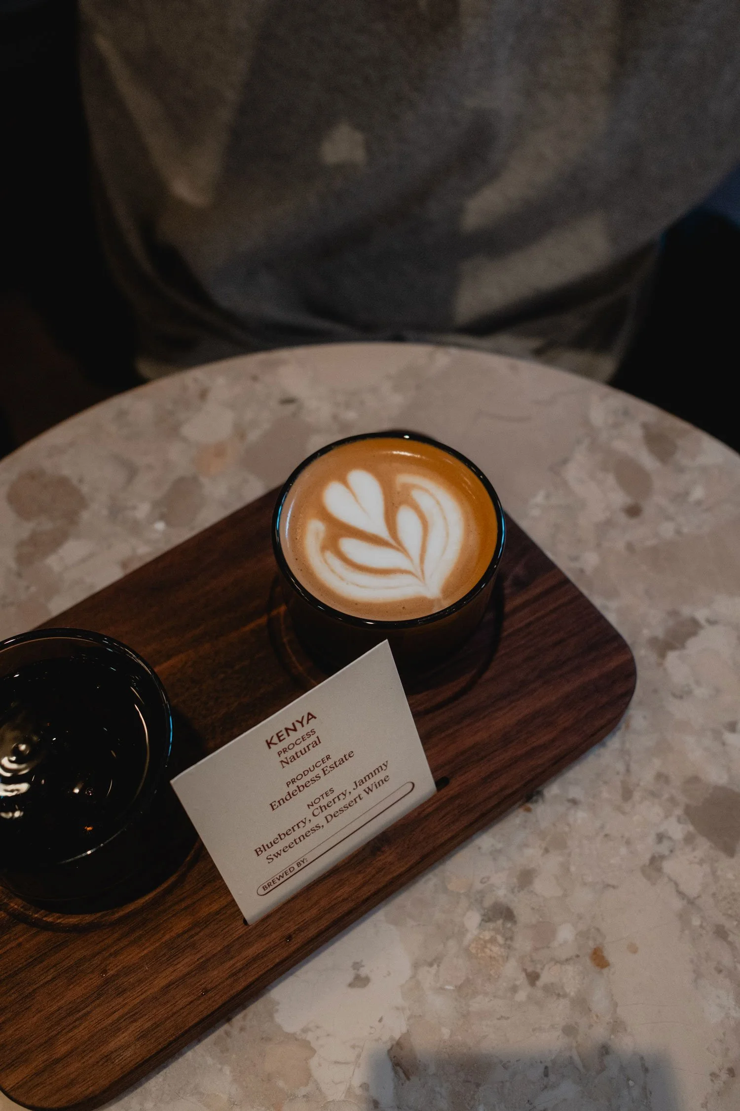 A latte with latte art on top, served on a wooden tray with a second drink, on a round marble table.