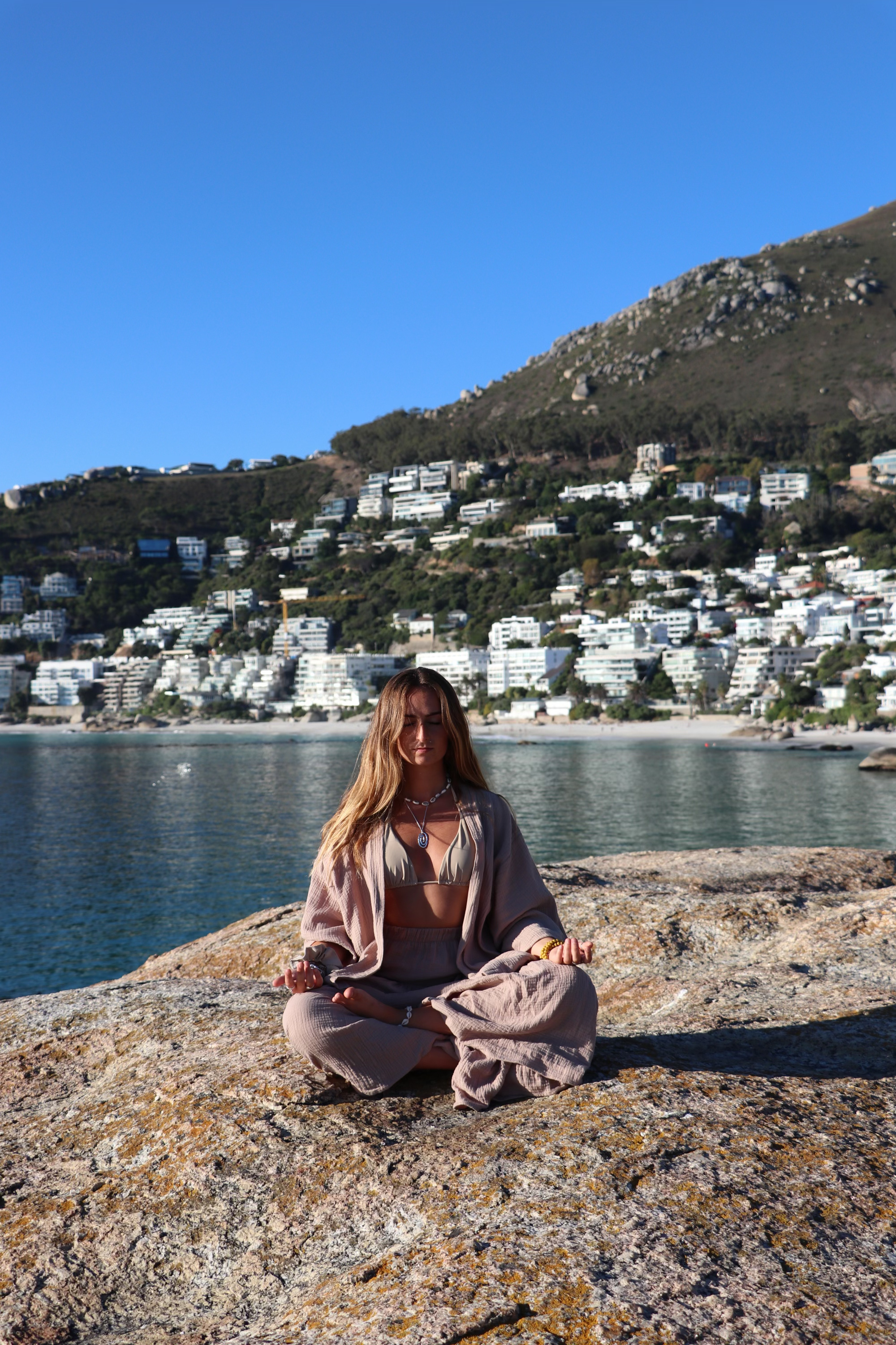 A woman with long hair meditating in a seated yoga pose on a large rock by the water, with a backdrop of a hillside cityscape and clear blue sky.