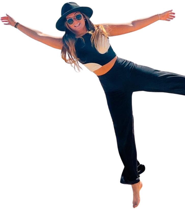 A woman wearing a black wide-brimmed hat, sunglasses, a black sleeveless top, and black pants, standing with arms outstretched and smiling.