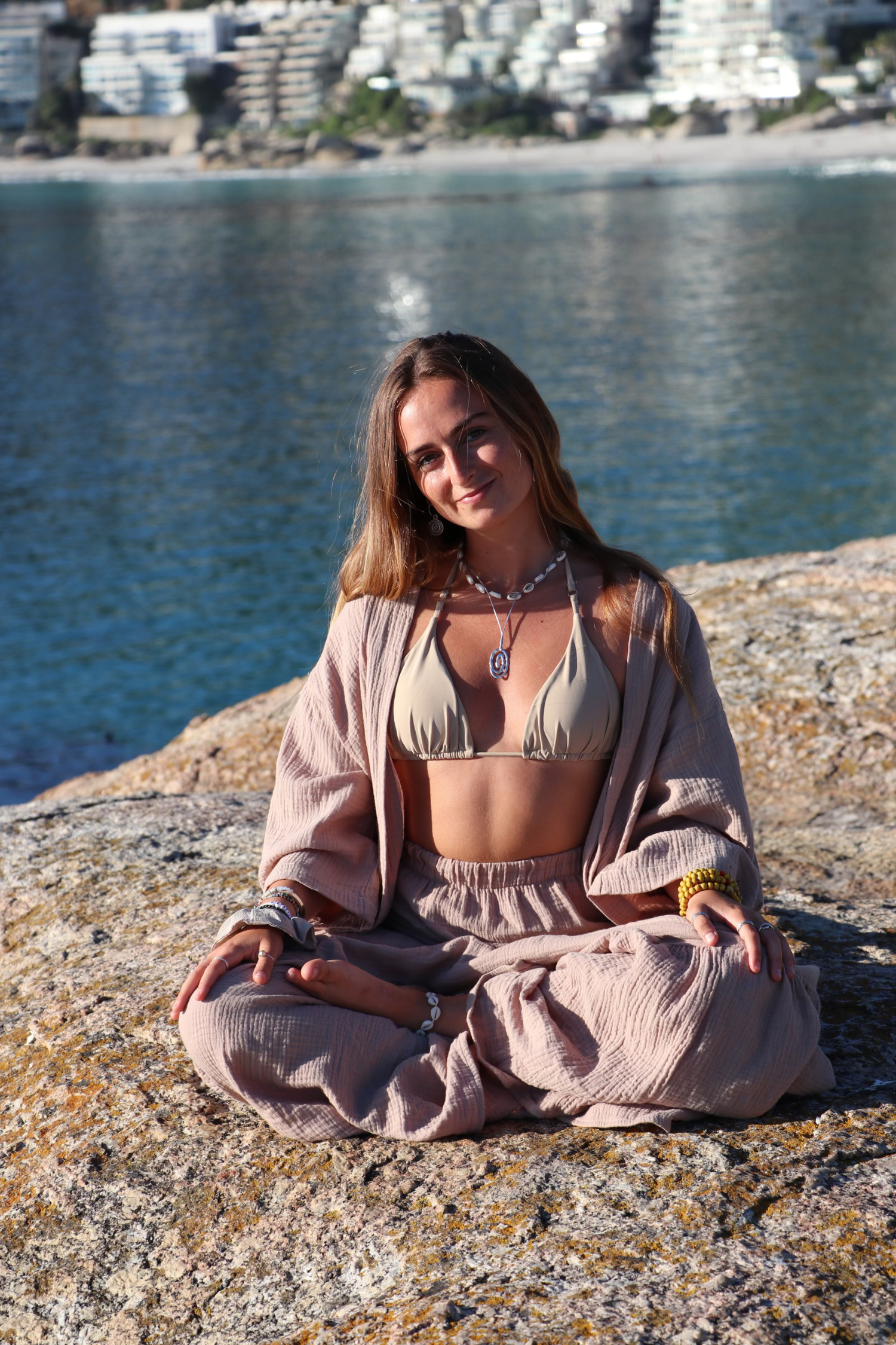 A woman sitting cross-legged on a large rock by the water, with a cityscape in the background, wearing bohemian-style clothing, necklaces, and bracelets.