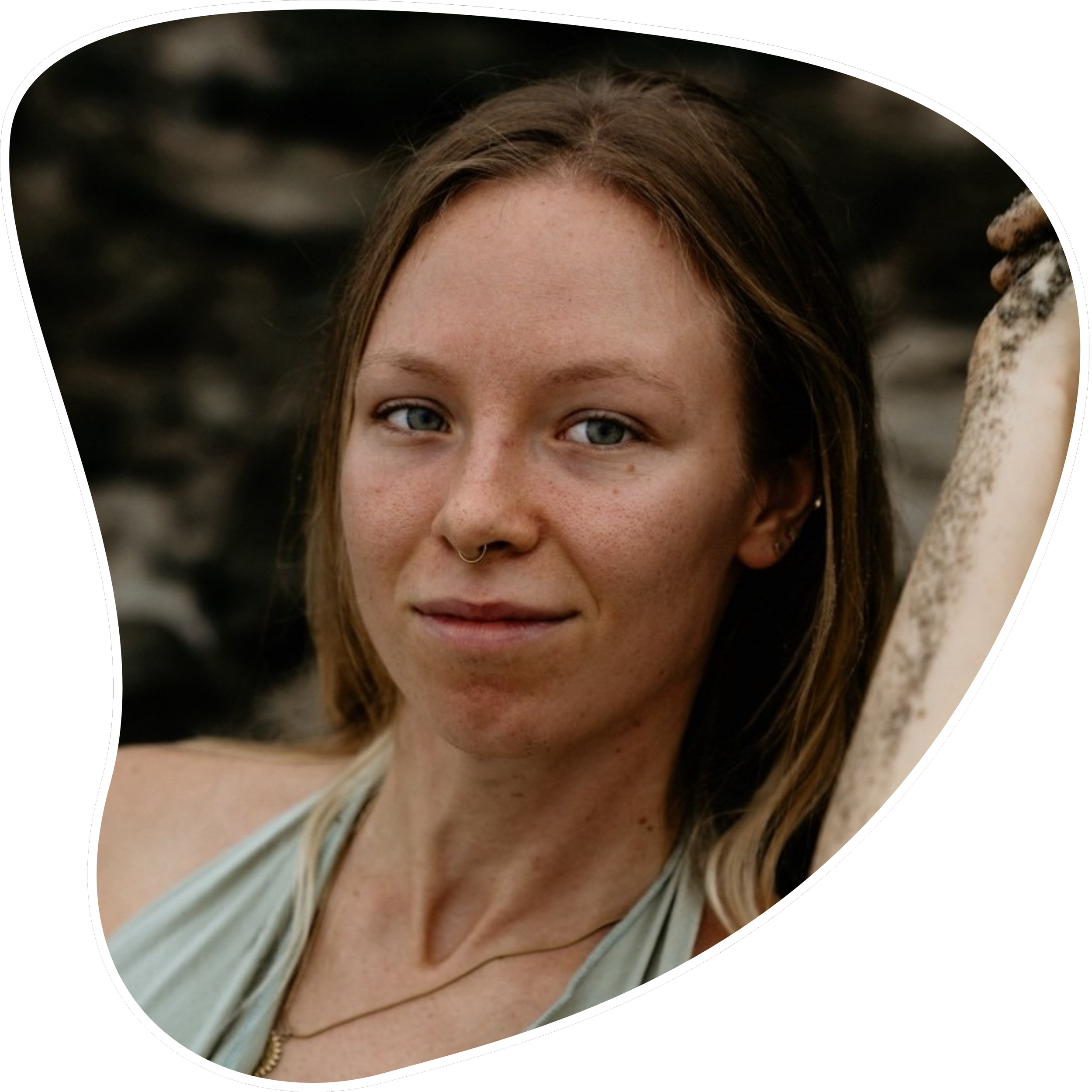 Close-up portrait of a young woman with brown hair, blue eyes, and a septum piercing, outdoors against a blurred natural background.