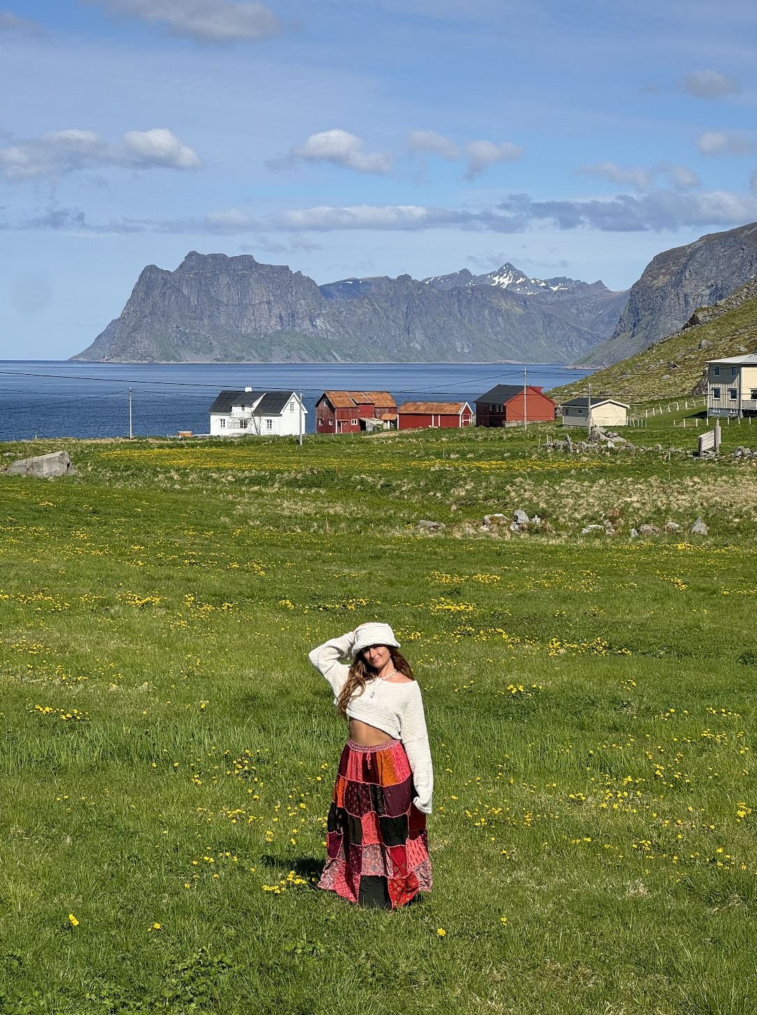 A woman standing in a grassy field with yellow flowers, wearing a white hat and a long, colorful skirt, with a mountainous landscape, houses, and water in the background.
