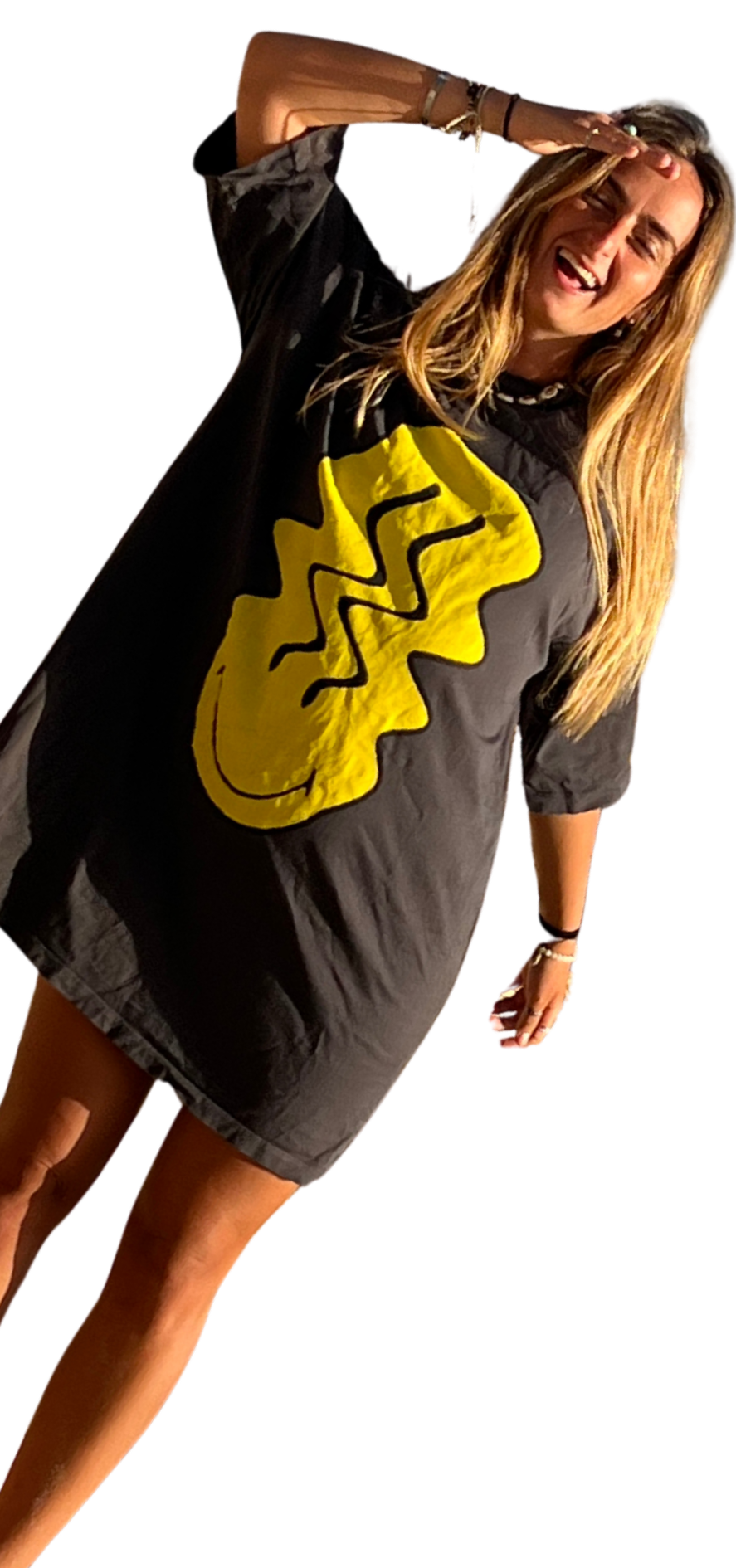 Woman with long blonde hair smiling and laughing, wearing a black oversized T-shirt with a large yellow smiley face with jagged lines on it, standing with one hand on her head and the other hanging down, against a black background.