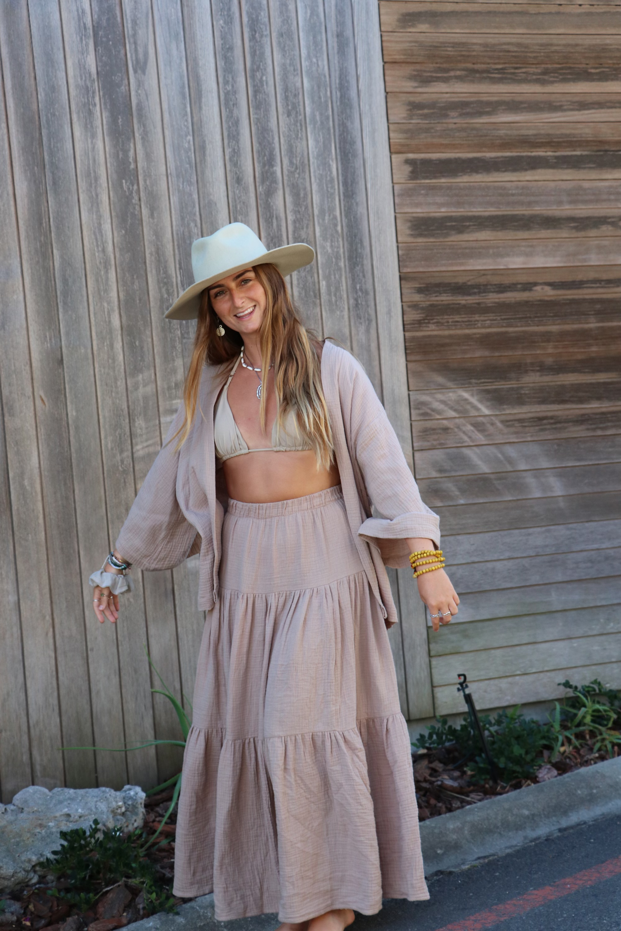 A woman with long hair wearing a wide-brimmed hat, beige bikini top, and a flowing beige skirt standing outdoors against a wooden fence.