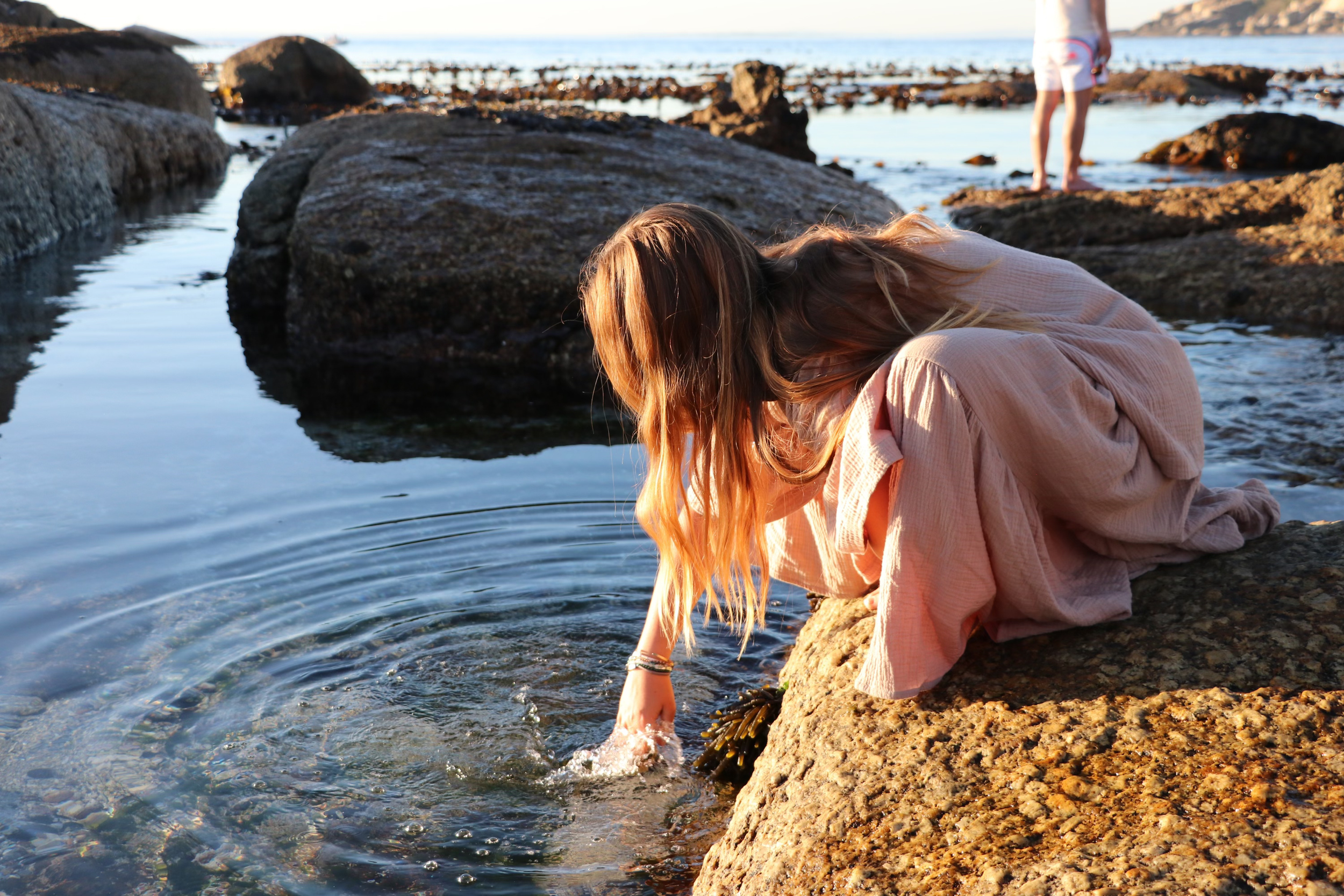 A woman with long hair crouching on rocks by the water, reaching into the pool of water with her hand, at the beach during sunset. In the background, a person stands on rocks near the water.