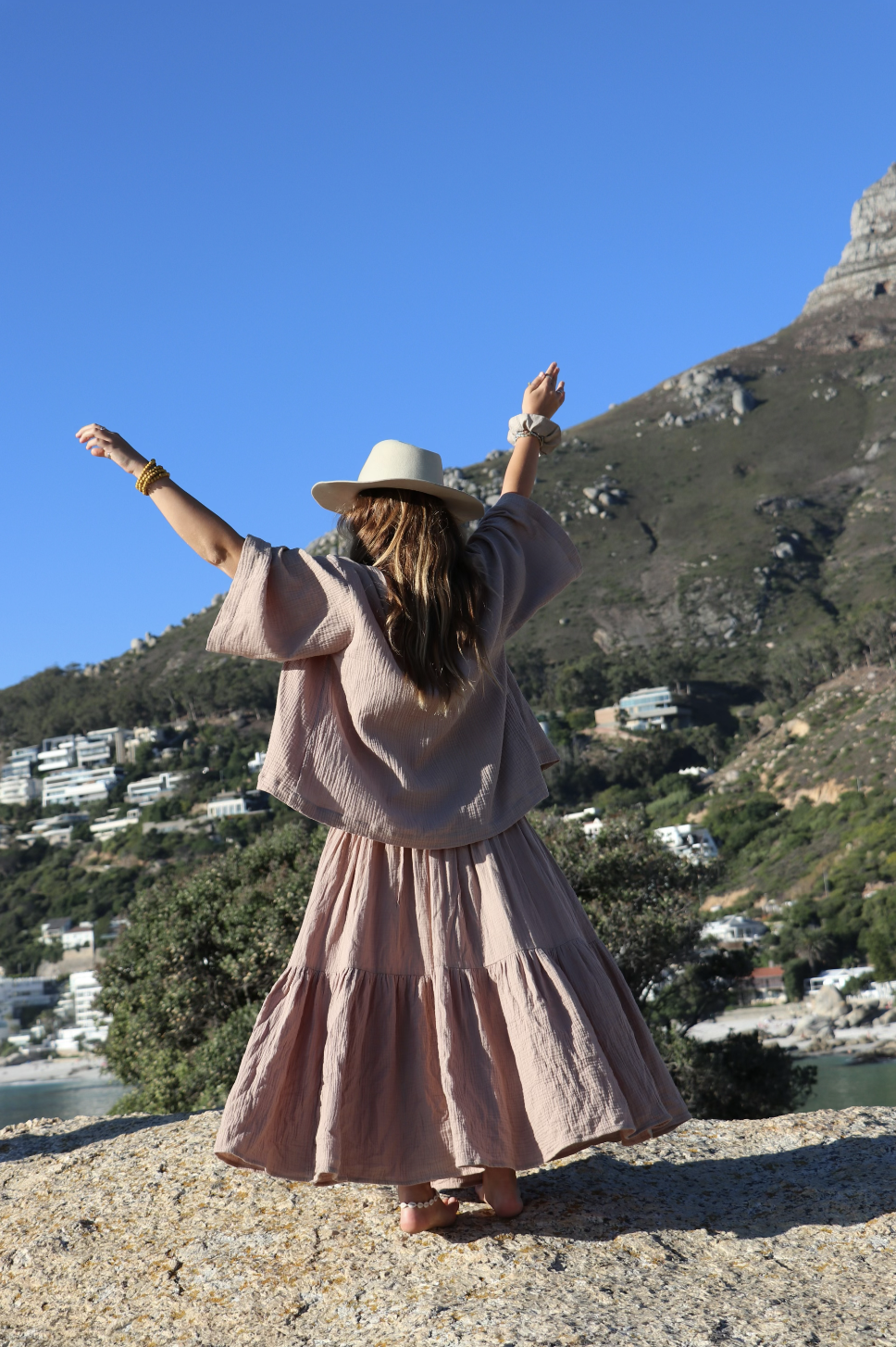 Woman in a beige outfit and hat standing on a rocky surface with arms raised, overlooking a hillside with houses and greenery under a clear blue sky.