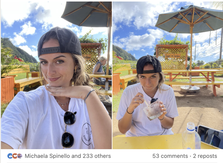 A young woman with long brown hair, wearing a black baseball cap, white t-shirt, and sunglasses hanging from her shirt, taking a selfie outdoors under a cloudy sky; in the background, another person wearing a white t-shirt and black cap sits at a yellow table, eating a snack, with a large umbrella and scenic landscape behind them.