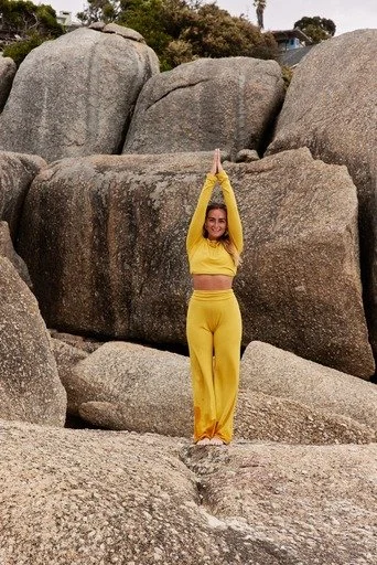 Woman in yellow workout clothes standing with arms raised, touching her hands together above her head, on a rocky landscape with large boulders.
