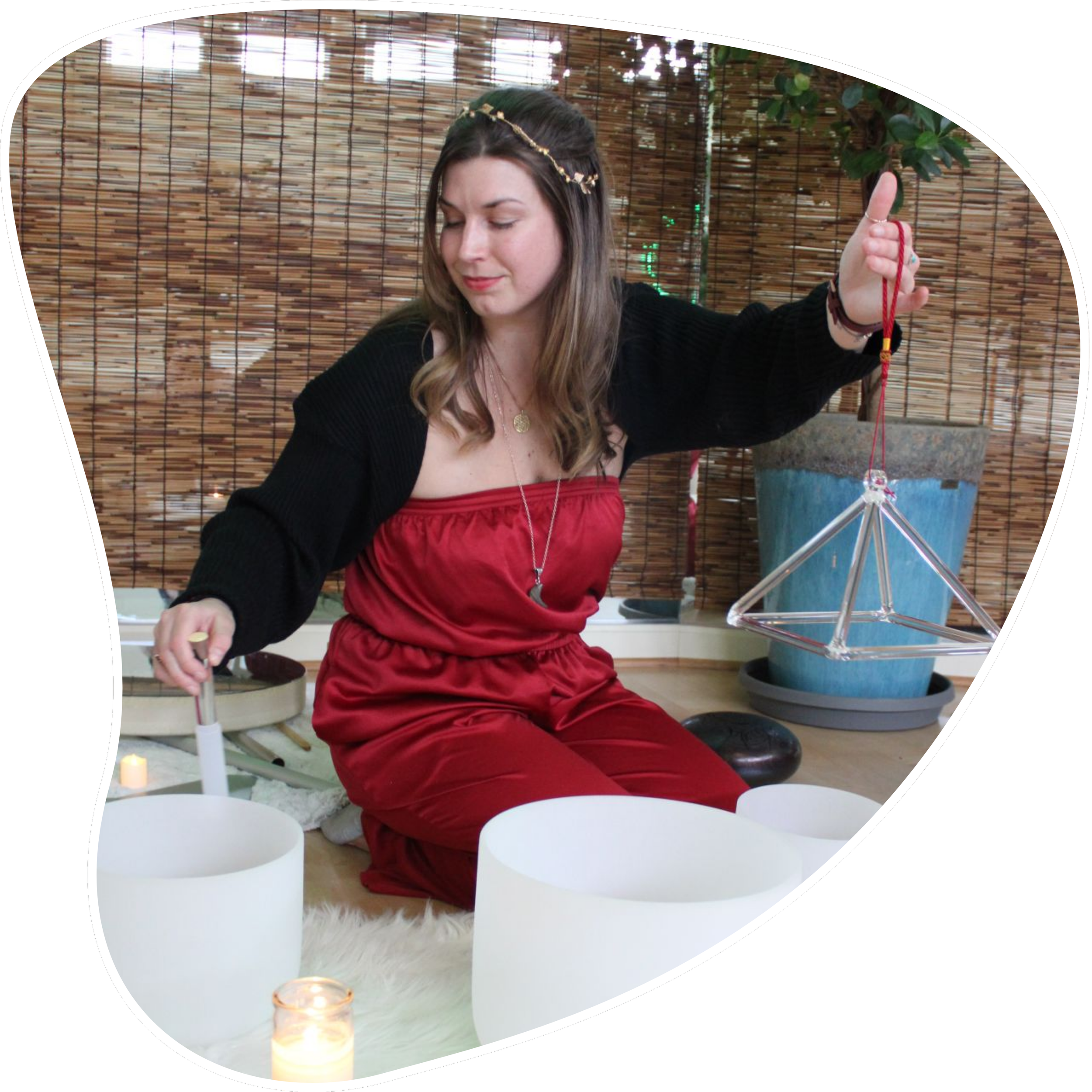 A young woman dressed in a red strapless jumpsuit and a black cardigan, sitting on a furry rug, appears to be playing crystal singing bowls and holding a crystal pyramid ornament, with a bamboo curtain and a potted plant in the background.