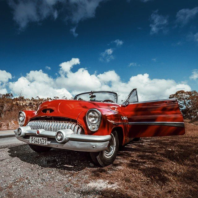 A vintage red convertible car with its door open, parked on a roadside under a partly cloudy sky.