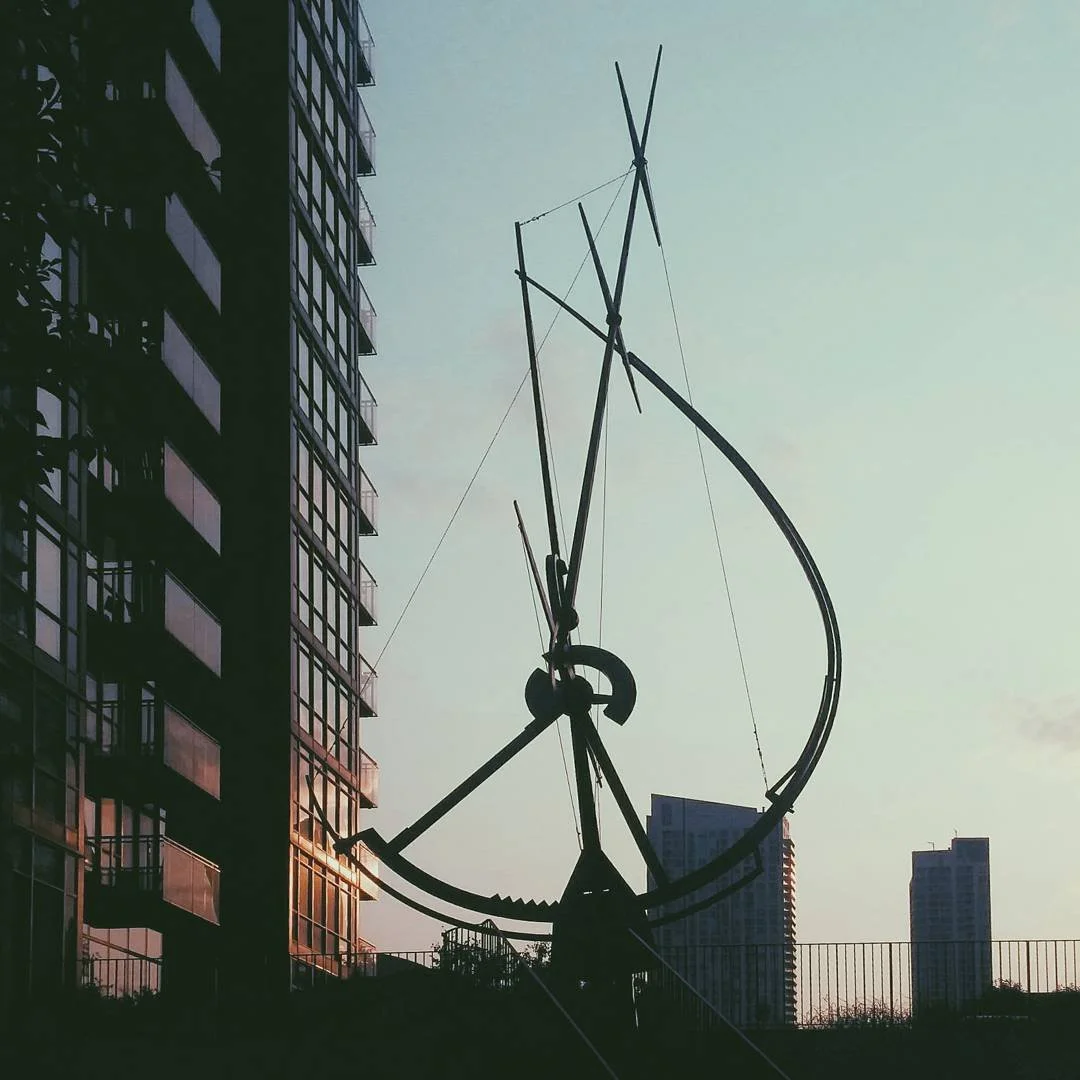 Silhouette of a modern outdoor sculpture resembling an abstract human figure against a sunset sky, with tall buildings in the background.