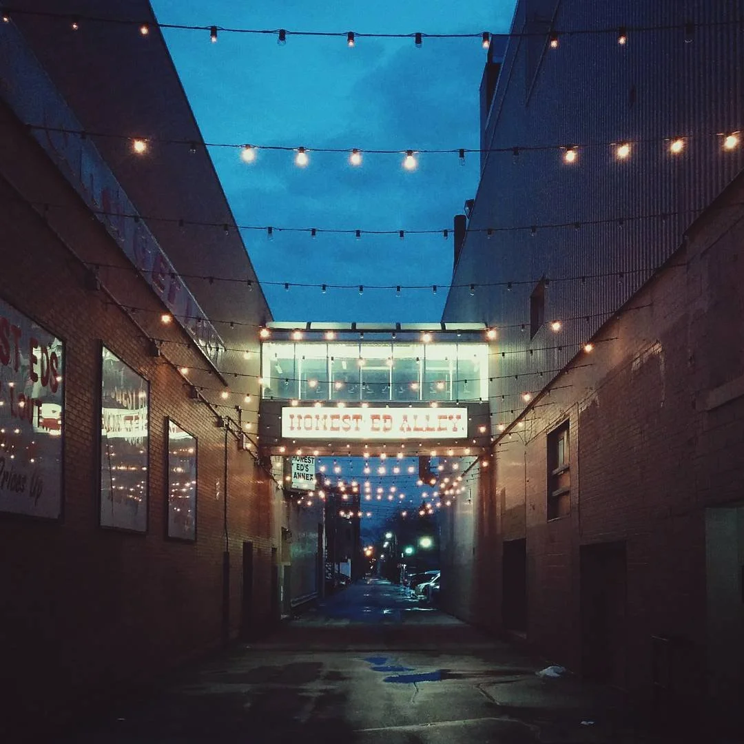 A dimly lit alley at dusk with string lights hanging overhead, leading to a glass building with a sign that reads 'Honest Ed's Alley'.