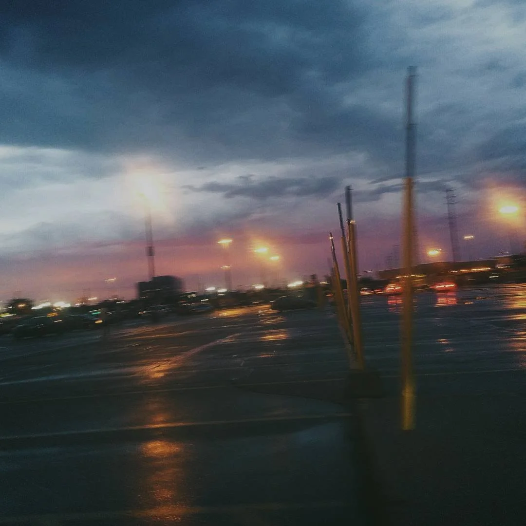 dusk at a parking lot with wet ground, illuminated by streetlights, with dark clouds in the sky and rain reflections on the surface.