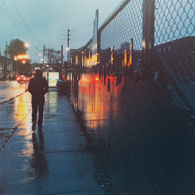 A person walking on a wet sidewalk near a fence, with streetlights and a bus in the background during dusk or dawn.