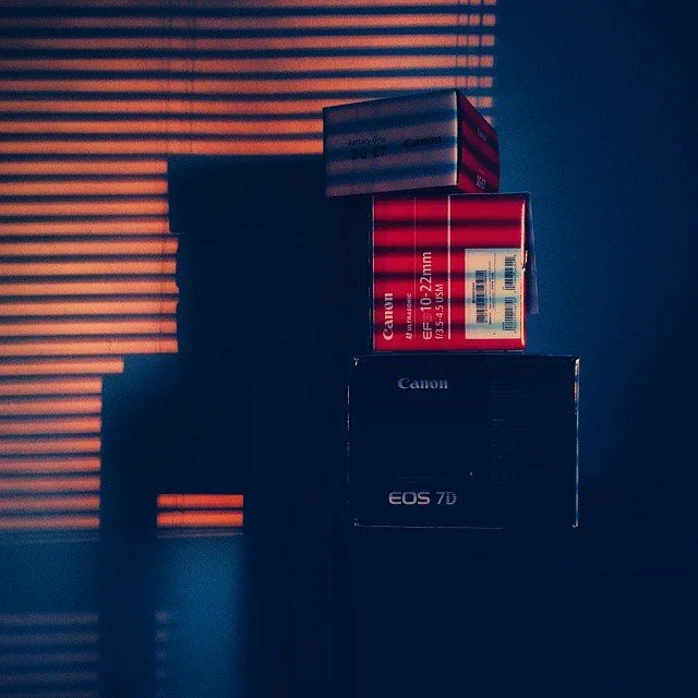Stacked boxes of camera equipment, including a Canon EOS 7D camera box and red and white boxes, with shadow and horizontal light streaks on the wall.