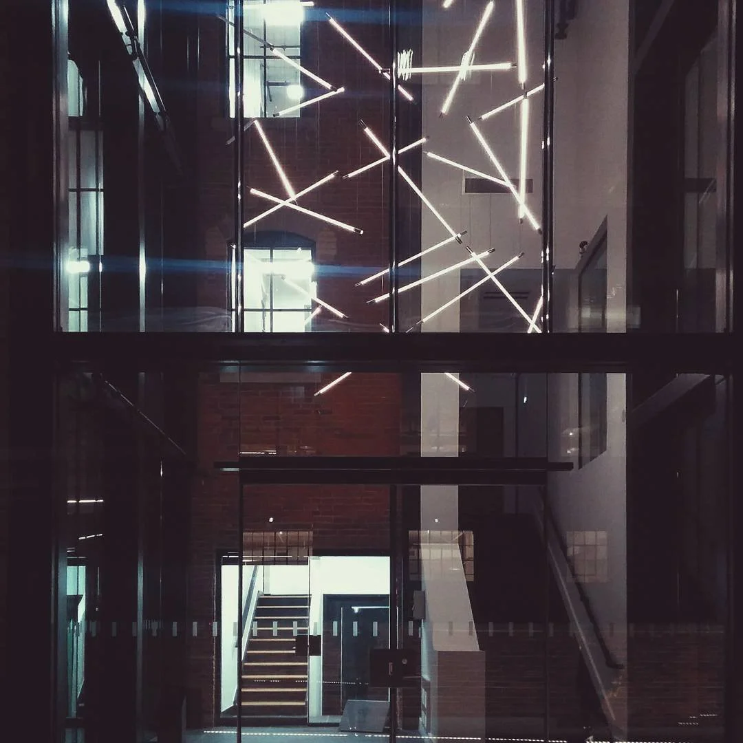 Interior view of a modern building with glass walls and an illuminated geometric light art installation on the ceiling.