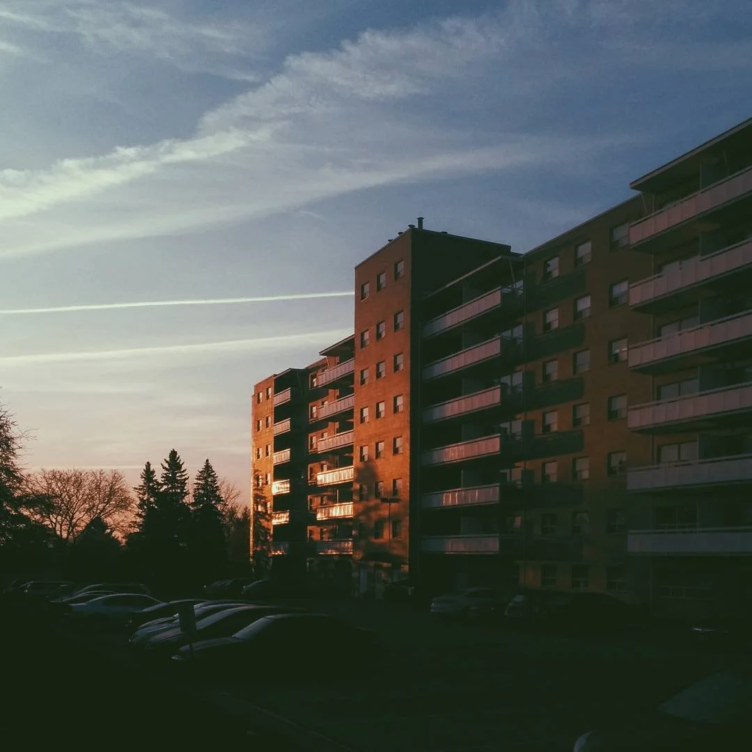 Sunset reflecting on a tall apartment building with a parking lot in the foreground, trees in the background, and a mostly clear sky with wispy clouds.