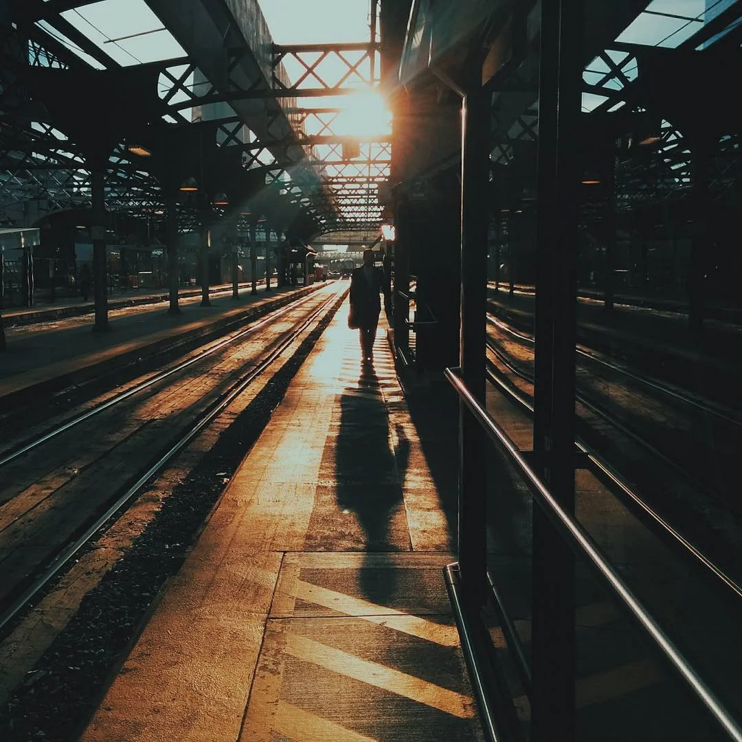 A person walking along a train platform during sunset, with their shadow cast on the ground, and train tracks running parallel to the platform.