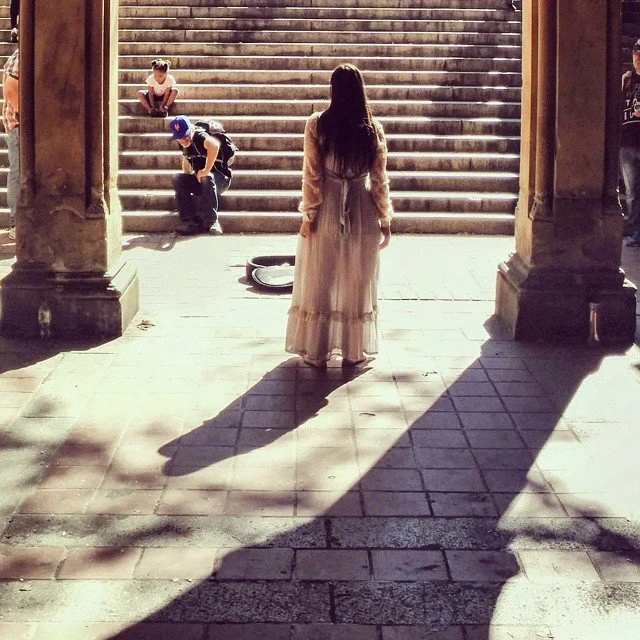 A woman in a long dress standing at the entrance of a building, casting a large shadow on the tiled floor, with two children on the steps behind her.