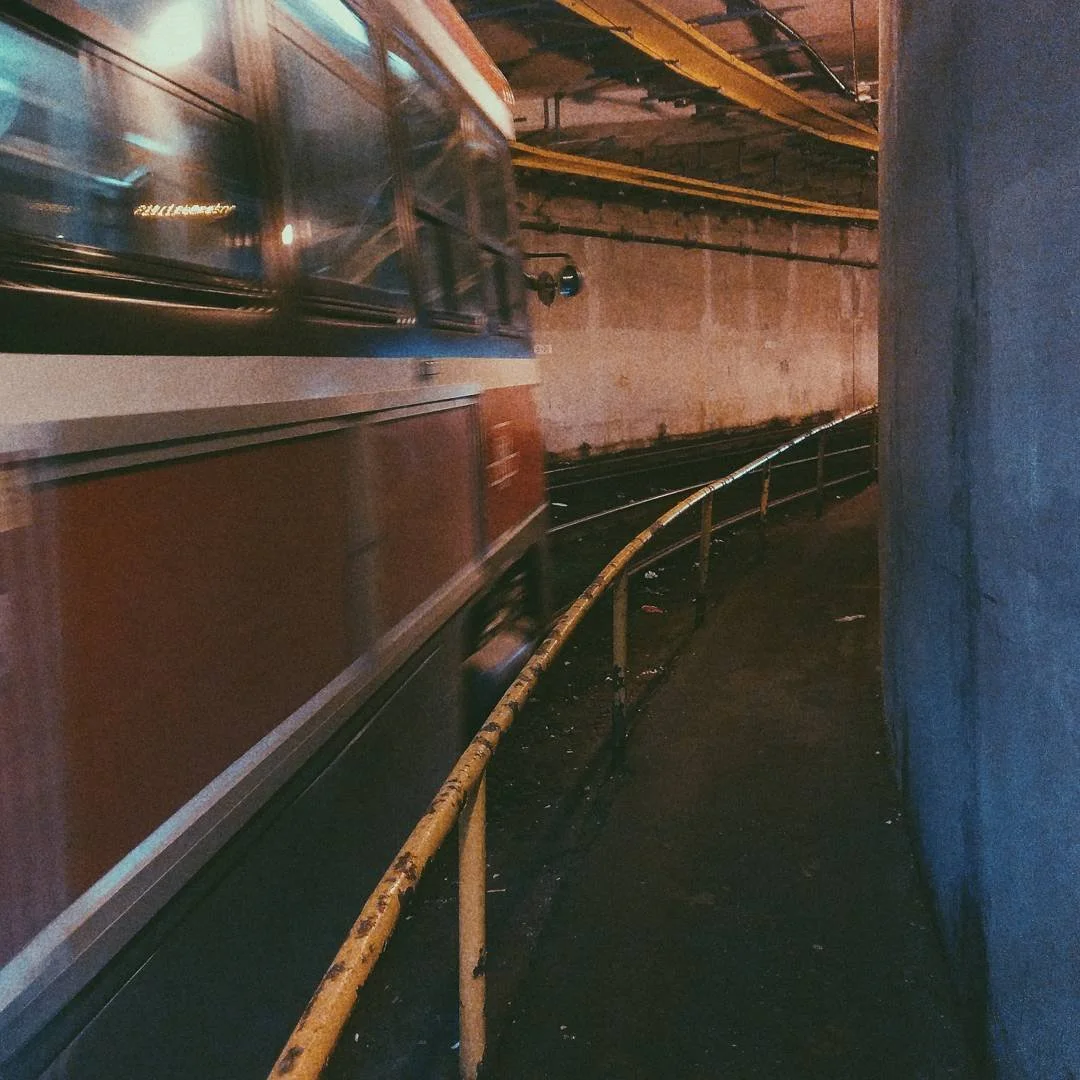 A moving train with red and silver exterior passing along an underground track, with a curved yellow safety railing and a concrete wall in the background.