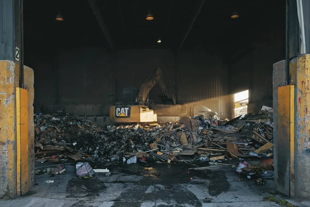 Inside a warehouse with a large pile of mixed debris and trash. A Caterpillar excavator is sorting the waste, with sunlight shining through an opening on the right side.