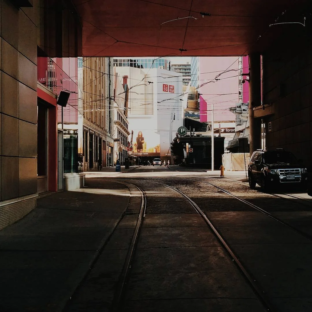 View of city street through a tunnel with tram tracks, parked cars, and tall buildings, including a UNIQLO billboard in the background.