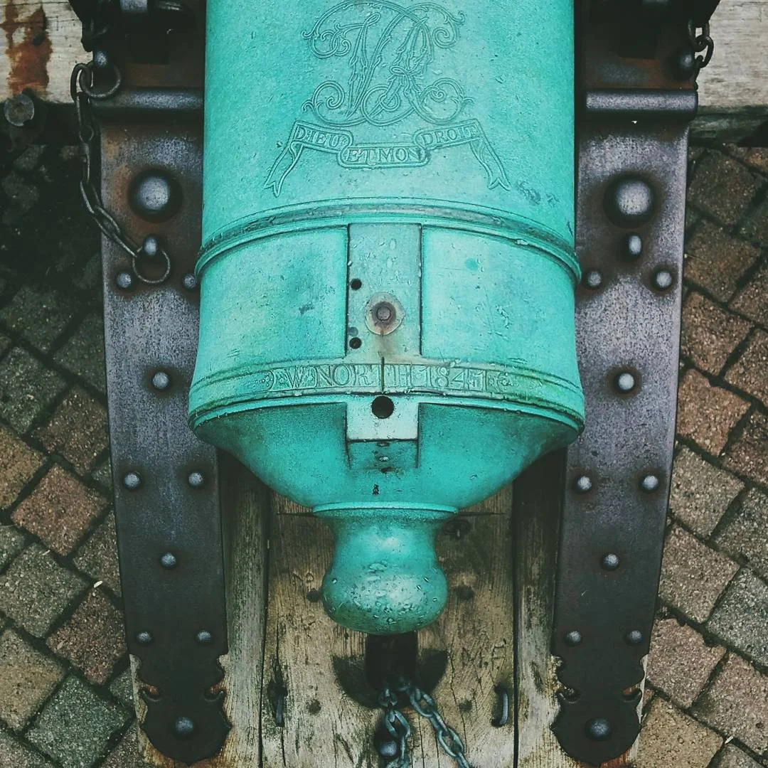 A vintage bronze cannon painted in turquoise, mounted on wooden and metal supports on a brick pavement.