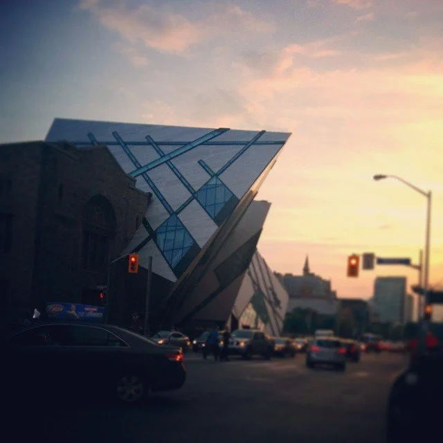 A modern building with an angular, geometric design and large glass windows in an urban setting during sunset, with traffic and streetlights.