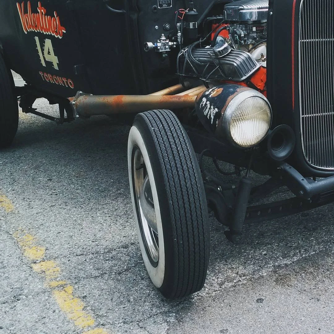 Close-up of a vintage race car showing the front left wheel, headlight, engine, and partial black body with red lettering that reads 'Valentine's 14 Toronto'; the car is on a textured asphalt surface.