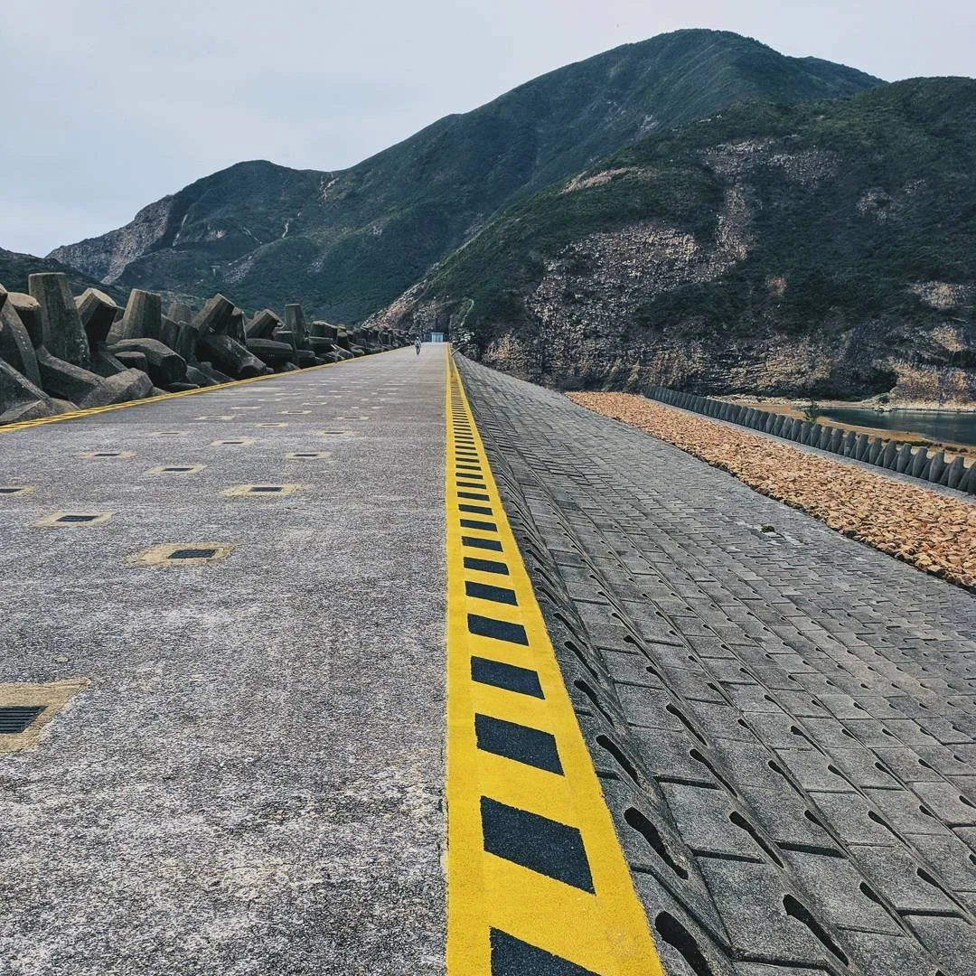 A long mountain road with a yellow and black striped border, surrounded by green hills and rocks on one side, with some concrete blocks on the other side.