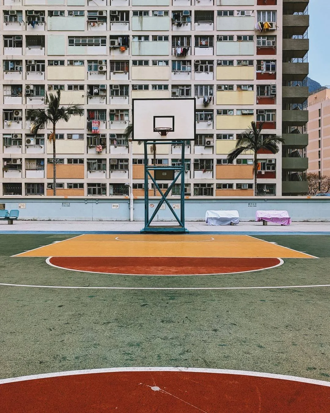 An empty outdoor basketball court with a building in the background, featuring colorful windows and balconies, two palm trees, and covered benches.