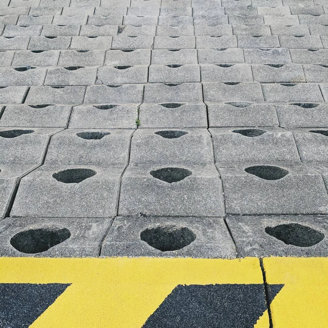 Concrete paving blocks with heart-shaped holes arranged in a grid pattern, with a yellow and black painted curb at the bottom of the image.