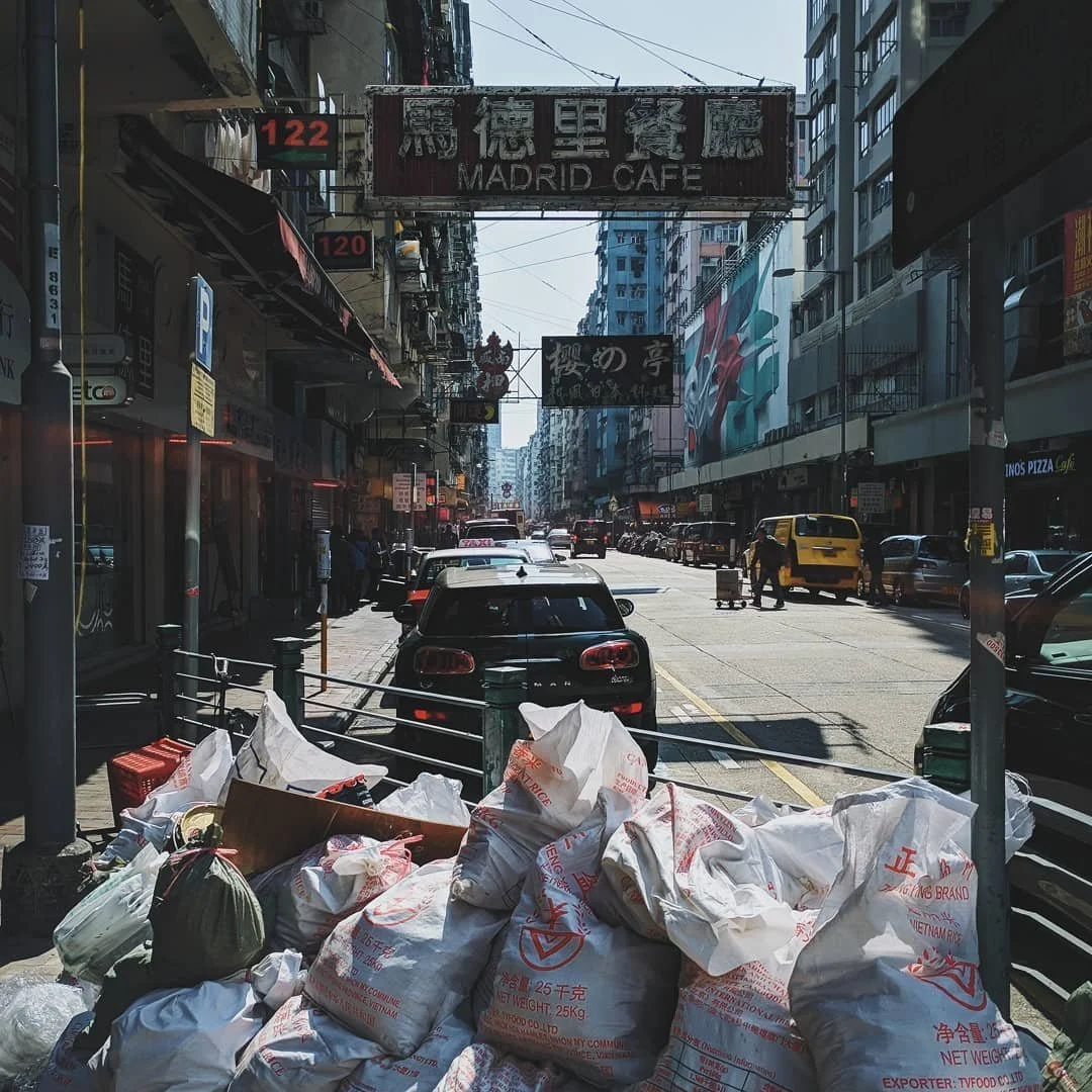 A busy city street with parked cars, taxis, and buildings with signs in various languages, including Japanese. There are bags and trash piled in the foreground.
