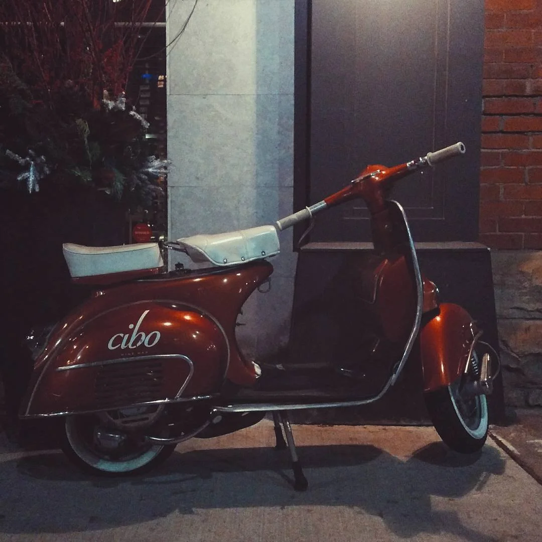 A vintage red scooter with a white seat and handlebar grips parked on the sidewalk at night, with a brick wall and greenery in the background.