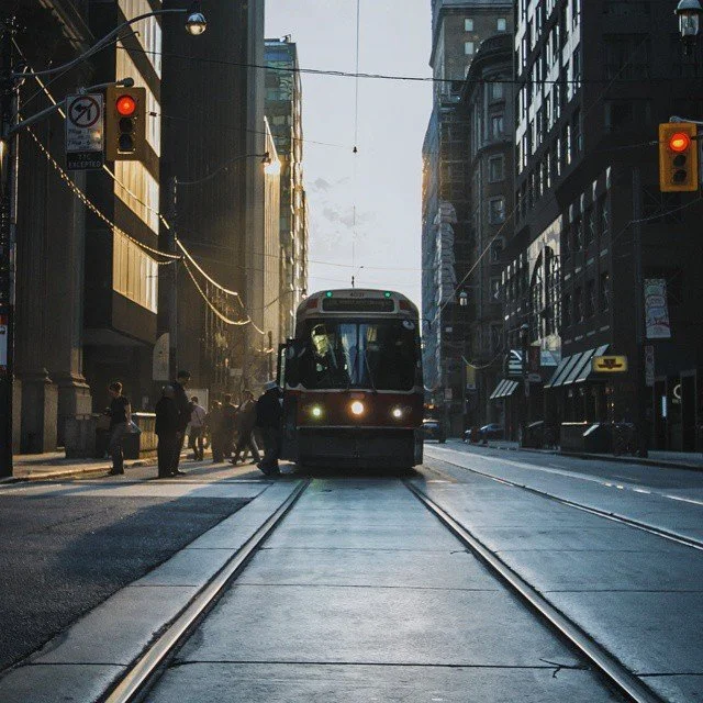 A city street with a tram approaching, pedestrians crossing, and traffic lights, bathed in early morning or late afternoon sunlight.