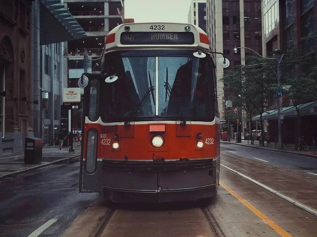 A streetcar or tram on city tracks in an urban area with tall buildings, trees, and pedestrians visible.