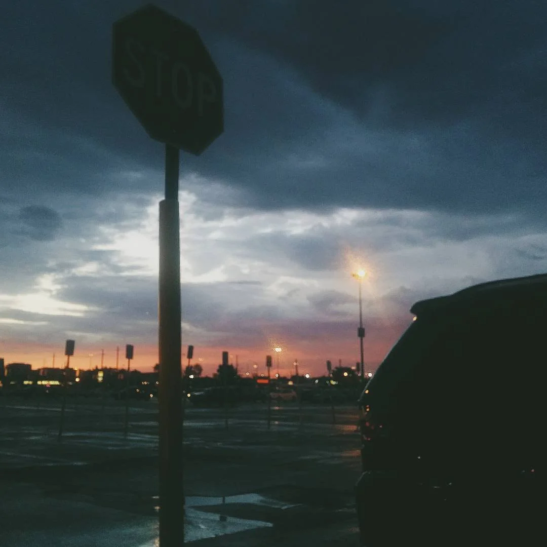 A stop sign in a parking lot during a sunset, with dark clouds in the sky and streetlights illuminated.