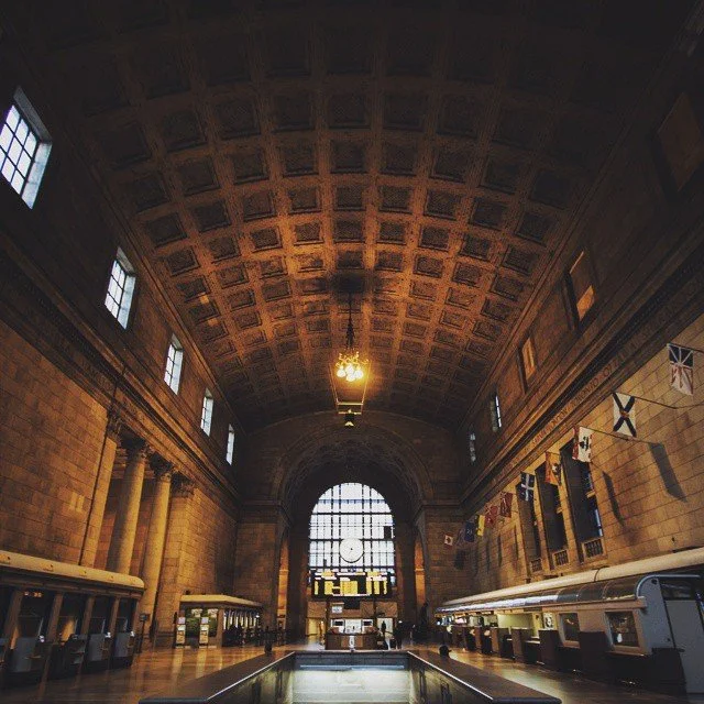 Interior of a historic train station with high arched ceiling, large windows, and a central clock above the main entrance.